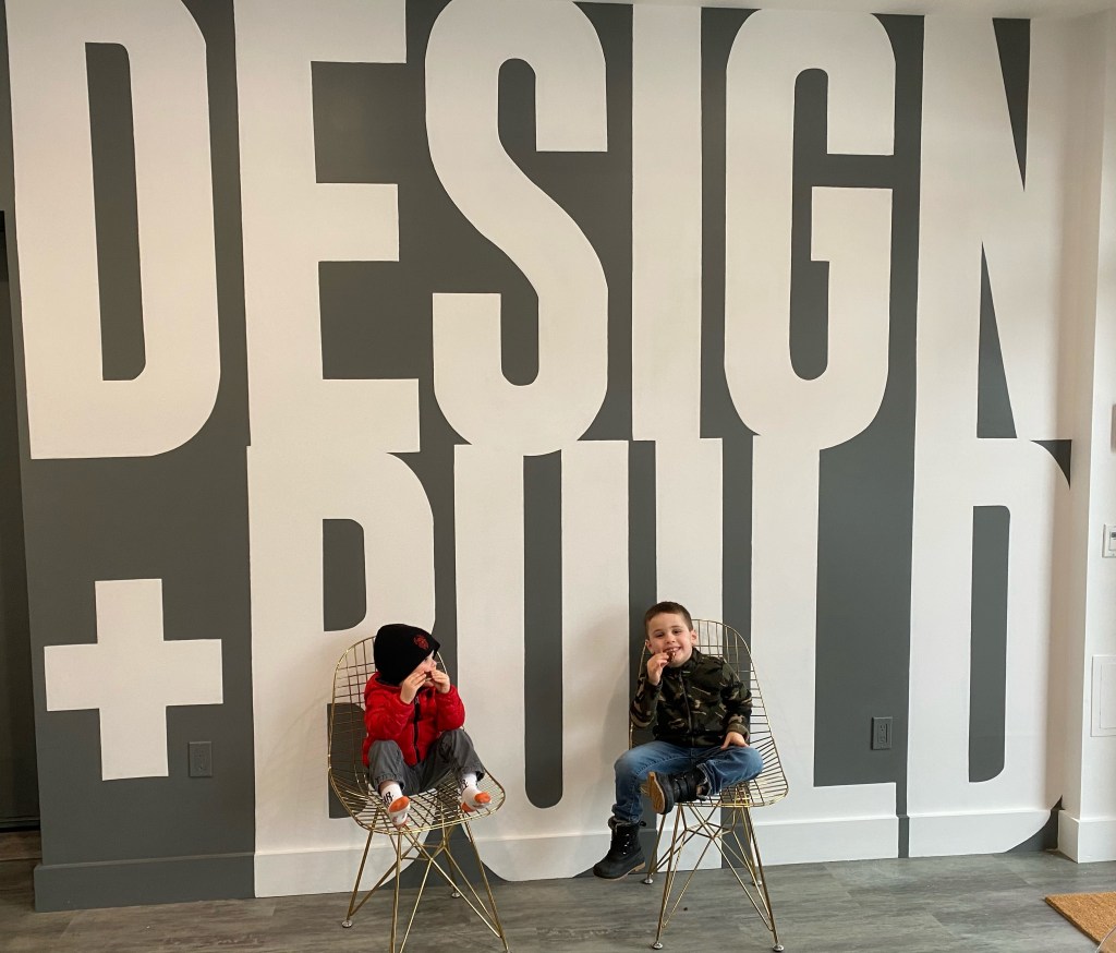 Mike's sons, THE Blette Brothers, sit in the company office with a mural "Design + Build" behind them.